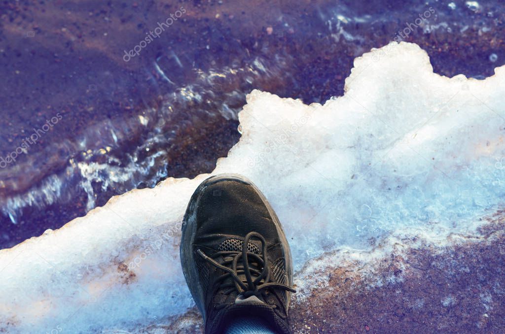 Person's foot wearing a trainer on melting ice near the lake.