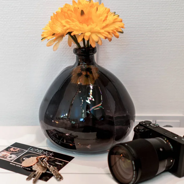 Black Shapeless Handmade Glass Vase with Yellow Flower in Back of a Sideboard with Postcard and Camera