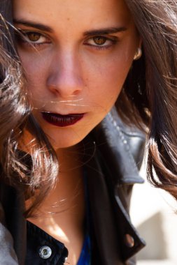 Güneşli bir günde, deri ceketli genç bir kadın, yoğun bakışlı, çok yakın çerçeveli.