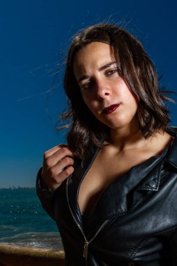 Arka planda deniz ve mavi gökyüzü olan bir portre Deri ceketli genç bir kadının güneşli ve rüzgarlı bir gününde