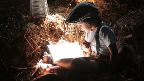 Copil dulce, băiete, citind o carte la mansardă pe o casă, stând pe un fân de paie, mâncând rulouri de coacere — Videoclip de stoc