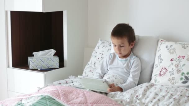 Больной мальчик лежит в постели с лихорадкой, отдыхает, играет на планшете — стоковое видео