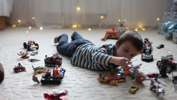 Niño pequeño jugando con un montón de juguetes de plástico de colores en el interior, la construcción de diferentes coches y objetos en Navidad — Vídeos de Stock