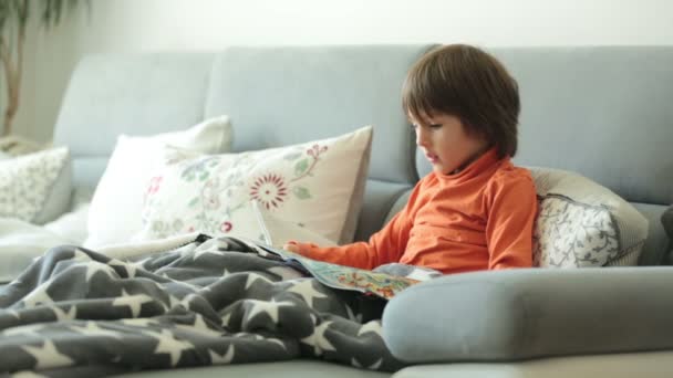 Niño, leyendo una revista, sentado en un sofá, cubierto con una manta suave — Vídeos de Stock