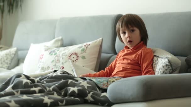 Kleiner Junge, liest eine Zeitschrift, sitzt auf einem Sofa, bedeckt mit weicher Decke — Stockvideo