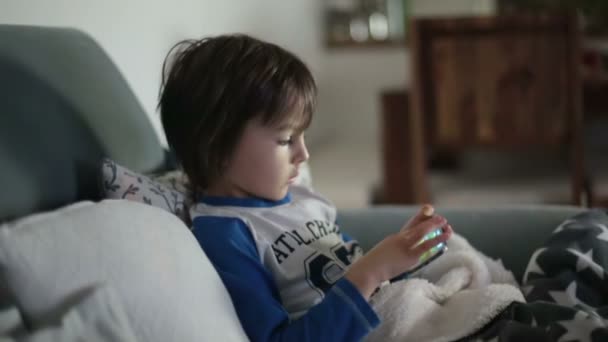 Pequeño niño preescolar, niño, jugando en el teléfono móvil en el sofá por la noche — Vídeos de Stock