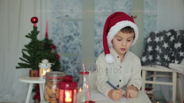 Vorschulkind, Junge, Brief an den Weihnachtsmann zu Hause schreiben, während es draußen schneit, Kekse essen — Stockvideo