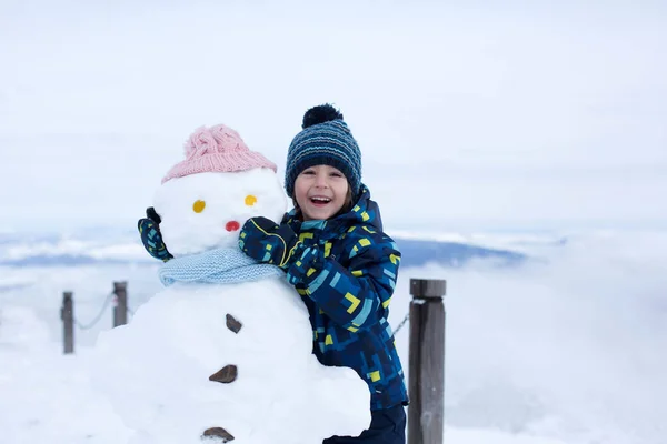 Милый ребенок, строит снеговика и играет с ним на вершине горы — стоковое фото