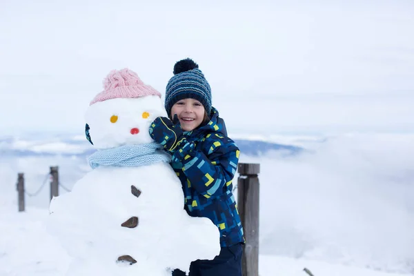 Милый ребенок, строит снеговика и играет с ним на вершине горы — стоковое фото
