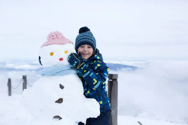 Criança bonito, construindo boneco de neve e brincando com ele no topo do monte — Fotografia de Stock