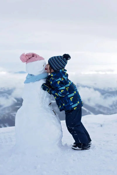 Criança bonito, construindo boneco de neve e brincando com ele no topo do monte — Fotografia de Stock