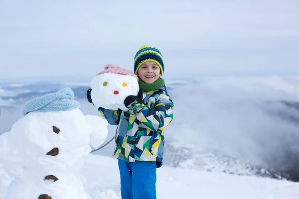 Criança bonito, construindo boneco de neve e brincando com ele no topo do monte — Fotografia de Stock