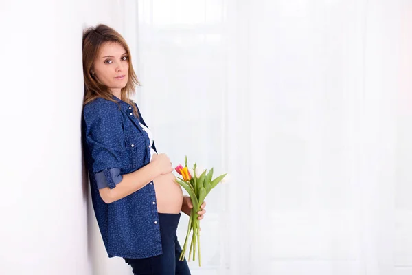 Mujer embarazada joven, sosteniendo flores de tulipán, apoyada en una pared — Foto de Stock