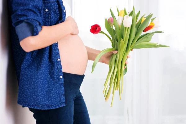 Mujer embarazada joven, sosteniendo flores de tulipán, apoyada en una pared —  Fotos de Stock