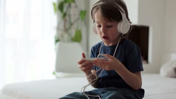 Симпатичный мальчик с телефоном и наушниками, слушающая музыка, в помещении, сидит на кровати в спальне — стоковое видео