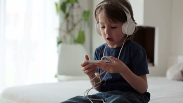 Lindo chico con teléfono y auriculares, escuchando música, en el interior, sentado en la cama en el dormitorio — Vídeos de Stock