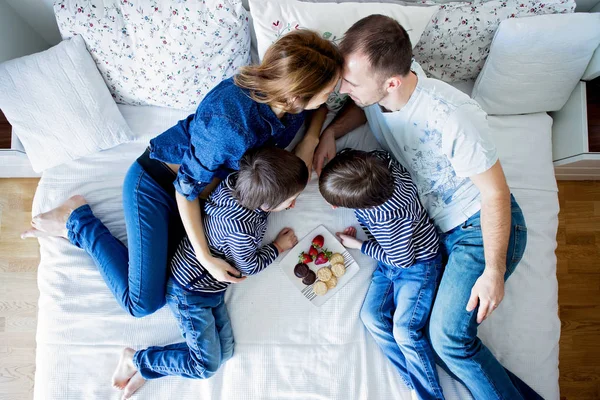 Schöne vierköpfige Familie, die auf dem Bett liegt und Erdbeeren isst — Stockfoto