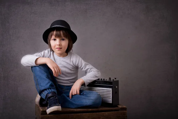 Netter Junge mit schwarzem Hut, sitzt auf einem Holztablett und hält ein Radio in der Hand — Stockfoto