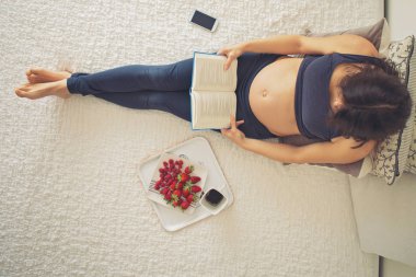 Genç hamile kadın, smartphone, kitap, kahve ile yatakta yalan