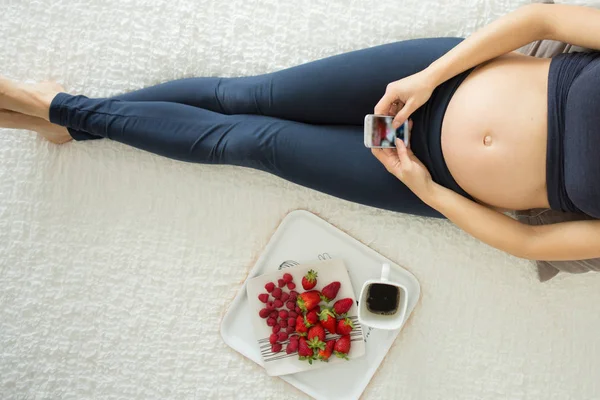 Jeune femme enceinte, couchée au lit avec smartphone, livre, café — Photo