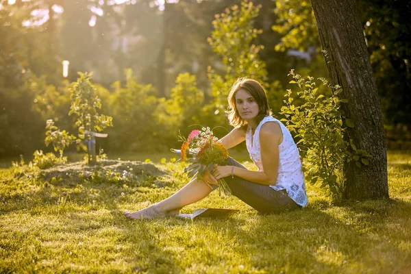 Genç kadın, bahçede oturan, buket çiçek tutan, kameraya, gün batımı yaz günü arıyorsunuz — Stok fotoğraf