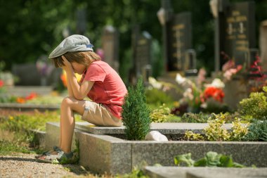 Üzgün bir mezarlığındaki mezar taşındaki oturan üzgün küçük çocuk,