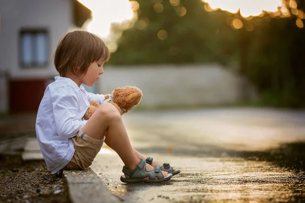 Triste niño, sentado en la calle bajo la lluvia, abrazando su t —  Fotos de Stock