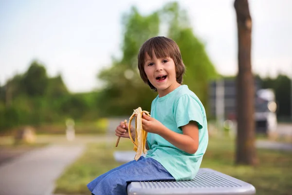 Aranyos gyermek, banán, ül egy padon, szabadtéri étkezési — Stock Fotó