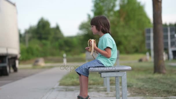 Χαριτωμένο παιδί, τρώει μπανάνα, ενώ κάθεται σε ένα παγκάκι υπαίθρια, άνοιξη — Αρχείο Βίντεο