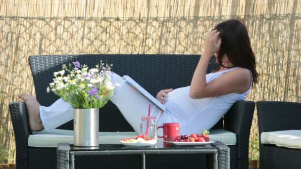 Young pregnant woman, having healthy breakfast, coffee, fruits and reading a book in a backyard garden — Stock Video
