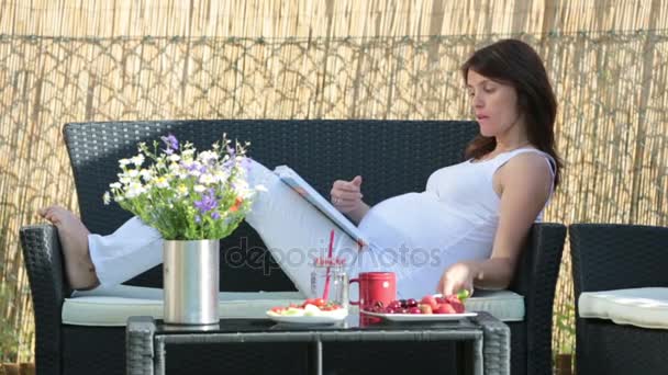 Mujer embarazada joven, desayunando sano, tomando café, frutas y leyendo un libro en un jardín del patio trasero — Vídeo de stock