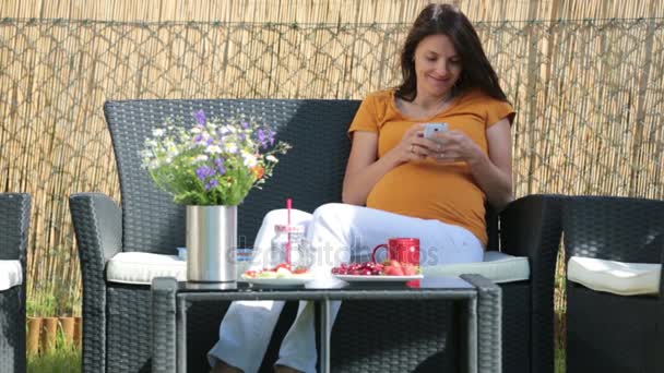 Mujer embarazada joven, desayunando sano, tomando café, frutas y leyendo un libro en un jardín del patio trasero — Vídeo de stock