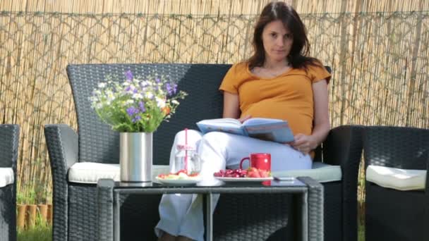 Young pregnant woman, having healthy breakfast, coffee, fruits and reading a book in a backyard garden — Stock Video