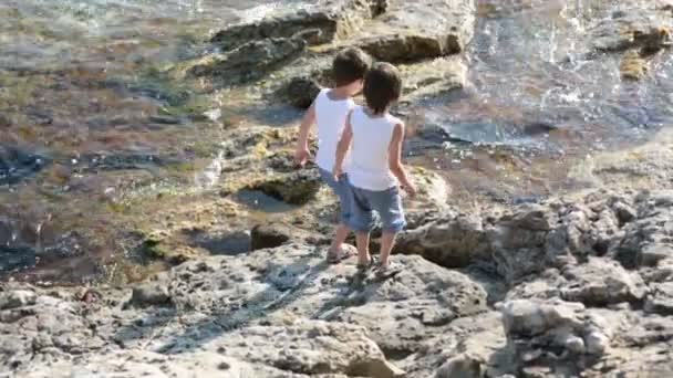 Dos niños, chicos, corriendo sobre rocas en la orilla del mar, divirtiéndose — Vídeos de Stock