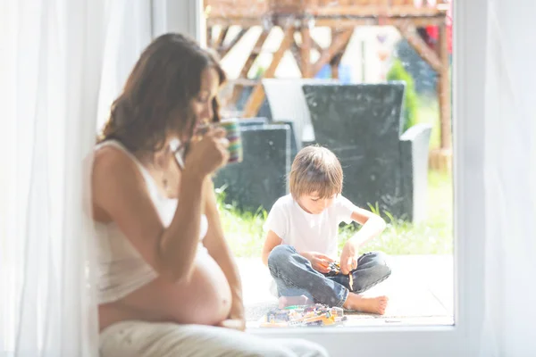 Молодая беременная женщина, сидя в комнате, наблюдая за своим ребенком — стоковое фото