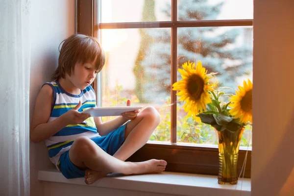 Lindo niño pequeño, jugando en la tableta en una ventana —  Fotos de Stock