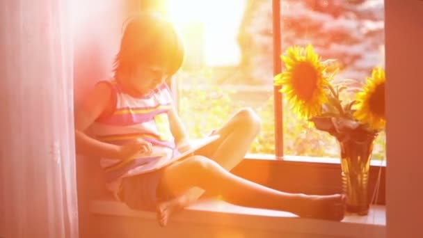 Criança pequena bonito, jogando no tablet em uma janela no pôr do sol — Vídeo de Stock
