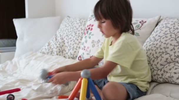 Сладкий мальчик дошкольного возраста, играющий с игрушками в помещении, сидя в кровати — стоковое видео