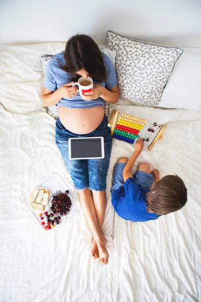 Mladá těhotná žena a její dítě, ležící v posteli s tabletem — Stock fotografie