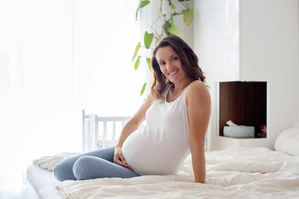 Feliz joven hermosa mujer embarazada, sentado en la cama en el dormitorio —  Fotos de Stock
