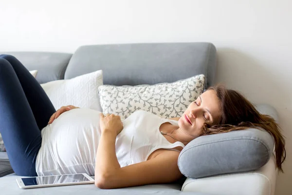 Νεαρή έγκυος γυναίκα, ξαπλωμένη στον καναπέ στο σαλόνι, αυτών — Φωτογραφία Αρχείου