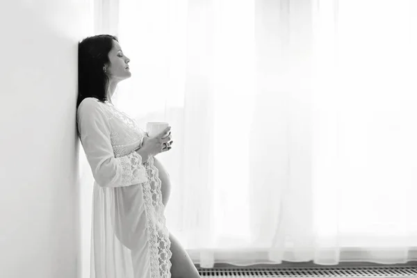 Beautiful young mom to be, leaning on a wall, having her coffee