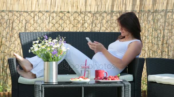 Young pregnant woman, having healthy breakfast, coffee, fruits and reading a book in a backyard garden — Stock Video