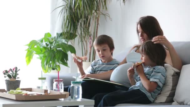 Joyeux jeune famille, mère enceinte et deux garçons, manger une délicieuse pizza à la maison, assis sur le canapé, lire un livre et rire — Video