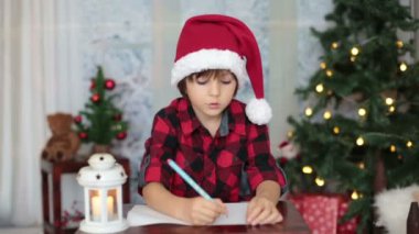 Sevimli küçük çocuk Noel dekorasyonu çevresindeki evde, Noel Baba'ya mektup yazma ahşap bir masaya oturup,