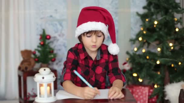 Ładny mały chłopiec, siedząc na drewnianym stole, pisząc list do Świętego Mikołaja w domu Bożego Narodzenia ozdoba wokół niego — Wideo stockowe
