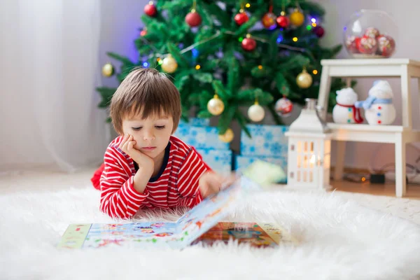 Χαριτωμένο αγόρι, διαβάζοντας ένα βιβλίο μπροστά από το χριστουγεννιάτικο δέντρο — Φωτογραφία Αρχείου