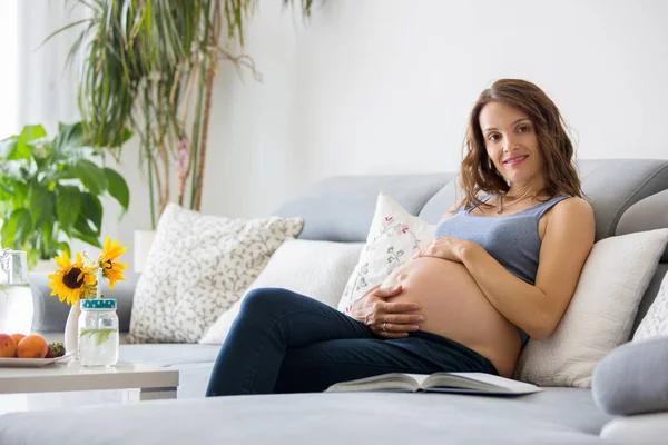 Fiatal szép terhes nő könyvével otthon a kanapén — Stock Fotó