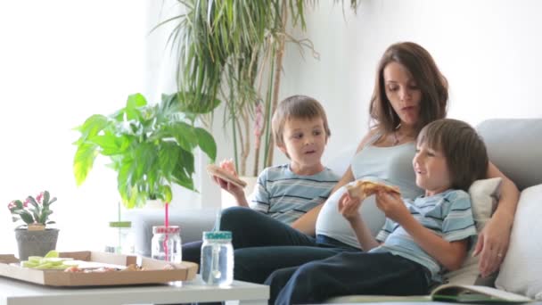 幸せな若い家族 妊娠中の母親と二人の少年 おいしいピザを家で食べる ソファーに座って 本を読んで 笑いを持っています — ストック動画