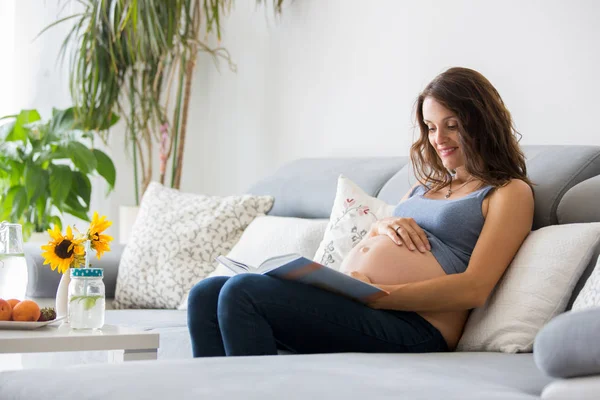 Mladá krásná těhotná žena čte knihu doma na gauči — Stock fotografie